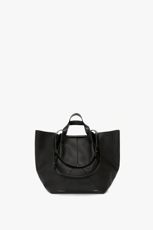 The **Victoria Beckham W11 Jumbo Tote In Black Leather** is a luxury leather tote in black with two sets of handles, one short and one long. The bag features a minimalist design, visible seams, and an internal pocket compartment for added convenience.