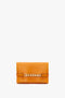 A rectangular bright mandarin leather clutch bag with a gold chain detail across the front and small text embossed at the bottom, Victoria Beckham UK Mini Chain Pouch Bag With Long Strap In Mandarin Leather.