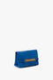 A sapphire blue rectangular clutch bag with a gold chain detail on the front. The words "Victoria Beckham UK" are embossed in gold on the lower right corner. This elegant piece, also known as a Mini Chain Pouch Bag With Long Strap In Sapphire Blue, features a detachable strap for versatile styling.