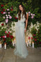 A woman stands in front of a lush floral backdrop with various flowers and candles. She is wearing a Gathered Tulle Detail Floor-Length Dress In Jade by Victoria Beckham and has long brown hair.