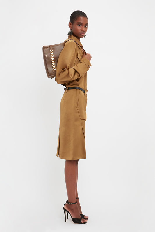A person stands sideways wearing a brown belted shirt dress and black high heels, carrying a khaki leather Chain Pouch Bag With Strap by Victoria Beckham UK over their shoulder.