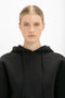 A person with blonde hair tied back is wearing a black Victoria Beckham Cropped Neoprene Hoodie In Black, posing against a white background.