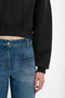 Close-up of a person wearing high-waisted blue jeans with front pockets and a Cropped Neoprene Hoodie In Black by Victoria Beckham, featuring a structured hood that highlights their midriff.