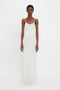 A black woman in a Victoria Beckham floor-length cami dress in ivory stands against a plain white background, looking directly at the camera.