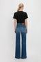 A person with a ponytail stands facing away, wearing a PARIS STREETS ARE MY RUNWAY T-shirt in Black and blue high-waisted wide-leg jeans, embodying the chic elegance of Victoria Beckham on the Paris streets as their runway.