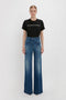 A person stands against a plain background wearing a Victoria Beckham PARIS STREETS ARE MY RUNWAY T-shirt in Black and blue high-waisted flared jeans with front patch pockets reminiscent of Victoria Beckham's iconic style.