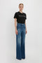 A person stands against a plain background wearing a black Victoria Beckham PARIS STREETS ARE MY RUNWAY T-shirt in Black paired with wide-leg blue jeans.