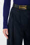 A person wearing a blue long-sleeve top, Victoria Beckham Wide Leg Trouser In Midnight, and a black belt with a gold buckle. Only the lower torso, hand, and a portion of the arm are visible.