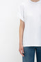 Person in an oversized Victoria Beckham Asymmetric Relaxed Fit T-Shirt In White with folded sleeves and blue jeans, standing against a blank white background. Only the upper body is visible.