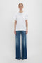 A person stands against a plain background wearing an oversized Victoria Beckham Asymmetric Relaxed Fit T-Shirt In White and blue wide-leg jeans.