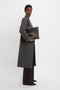 A person in a Victoria Beckham Tailored Slim Coat In Grey Melange and high-neck top holds a large, dark brown leather textured bag against a plain white background.