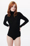 A person with long red hair wearing a black Victoria Beckham Poloneck Bodysuit In Black stands against a white background.