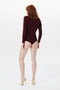 A person with red hair wearing a long-sleeved Poloneck Bodysuit In Bordeaux by Victoria Beckham and yellow high heels stands facing away from the camera against a white background.