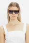 A woman with long blonde hair wears rectangular Victoria Beckham V Plaque Frame Sunglasses In Dark Brown Horn and a white sleeveless top against a white background.