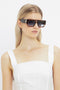 A woman with long blonde hair, wearing oversized rectangular V Plaque Frame Sunglasses In Dark Brown Horn from Victoria Beckham and a white sleeveless top, poses against a plain white background.