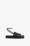 The Victoria Beckham Frances Sandal in Black features a black, open-toe design with a flat sole, a wide minimalist leather band over the foot, and an ankle strap with a buckle.