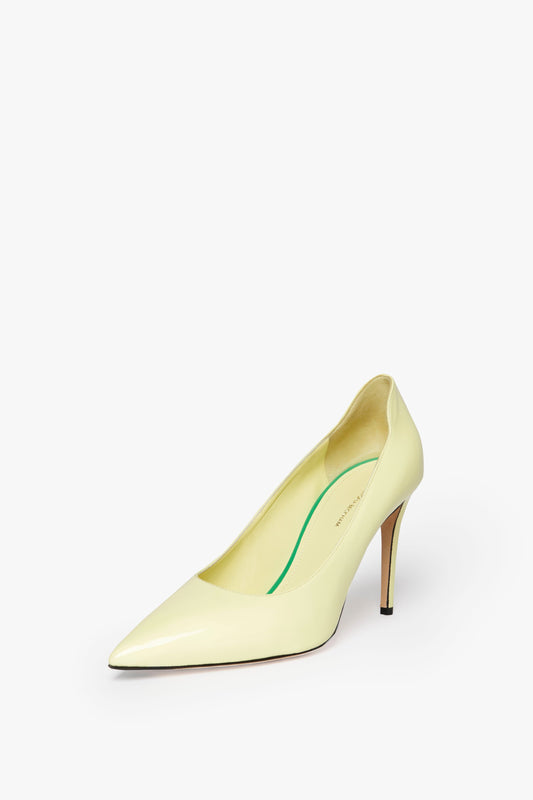 A single beige high-heeled shoe with a pointed toe, 90mm stiletto heel, and green inner sole on a white background. This VB 90mm Pump In Pale Lemon by Victoria Beckham is a true wardrobe essential.