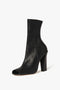 The Iona Boots In Black by Victoria Beckham is a black high-heeled ankle boot with a peep toe design, featuring a smooth lambskin leather finish and a sturdy sculptural heel.