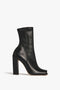 The Victoria Beckham Iona Boots In Black are a black, mid-calf leather beauty crafted from luxurious lambskin leather. They feature a high, sculptural block heel and a slim, pointed toe, all viewed elegantly from the side against a pristine white background.