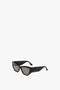 Black rectangular Sculptural Frame Sunglasses In Black with dark lenses and thick frames, crafted from glossy black acetate, displayed against a plain white background. The temples feature a small gold Victoria Beckham logo.