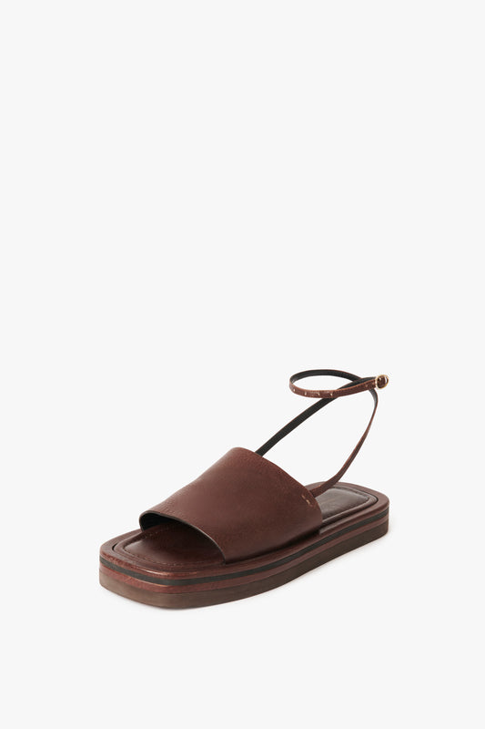 The Frances Sandal in Dark Brown by Victoria Beckham, boasting a 90s aesthetic, is a brown single-strap sandal with an ankle strap and thick leather and EVA sole, set against a white background.