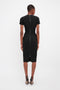 Person in Victoria Beckham's Fitted T-Shirt Dress In Black, short-sleeved, knee-length, with a high neck, back zipper and high heels, standing and facing away from the camera against a plain white background.