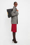 Person wearing a gray coat, red dress, patterned tights, and black high heels, carrying a large black bag over their shoulder. The bag is a stylish Jumbo Chain Pouch Bag In Black Leather by Victoria Beckham with Nappa leather and a detachable strap.