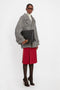 A person stands against a plain background, wearing a gray oversized coat, a white turtleneck, a red skirt, patterned tights, and high heels. They're holding the Jumbo Chain Pouch Bag In Black Leather by Victoria Beckham with its distinctive Nappa leather finish and detachable strap.