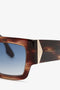 A close-up of V Plaque Frame Sunglasses in Dark Brown Horn with blue gradient, sculpted lenses and a gold triangular detail near the hinge. The Victoria Beckham brand name is visible on the arm.