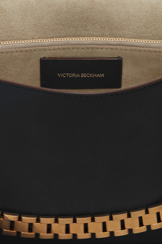 Close-up of an open Chain Pouch with Strap In Black Leather crafted from Nappa leather, displaying a suede interior and a zippered pocket with a label that reads "Victoria Beckham." The bag features a decorative strap with gold accents, offering versatile styling for any occasion.