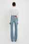 A person wearing a Victoria Beckham Cropped Patch Pocket Shirt In White and blue jeans stands facing away from the camera against a plain white background.