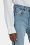 Close-up of a person wearing Victoria Beckham's Cropped Patch Pocket Shirt In White and blue jeans, focusing on the hand, shirt cuff, and waistband of the jeans.