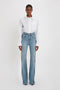 A person stands against a white background wearing a Victoria Beckham Cropped Patch Pocket Shirt In White paired with high-waisted blue jeans.