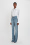 A person stands in front of a white background, wearing a Victoria Beckham Cropped Patch Pocket Shirt In White and high-waisted blue jeans with a visible seam design.