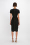 A person stands with their back to the camera, wearing a Spiral Fitted T-Shirt Dress In Black by Victoria Beckham, featuring a matte bonded crepe texture and a visible zipper down the back. Patterned high-heeled sandals complete the chic ensemble.