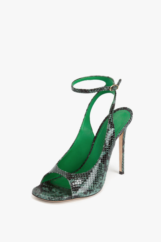 The Victoria Beckham Dawn Sandal in Petrol Green Printed Snakeskin is a high-heeled shoe with a striking green and black snake print, featuring an open toe, ankle strap with buckle, and green lining.