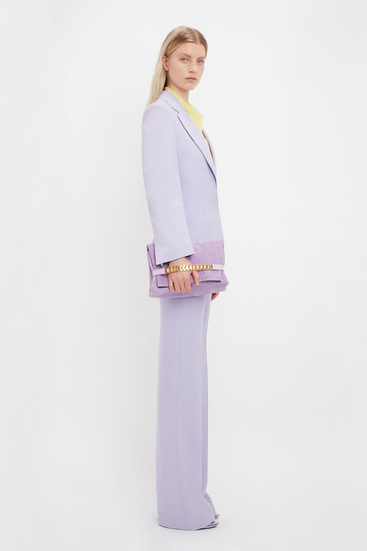 A woman with long blonde hair stands against a white background, dressed in a lavender suit, holding a Victoria Beckham Chain Pouch with Strap in Lilac Suede that matches her ensemble perfectly.