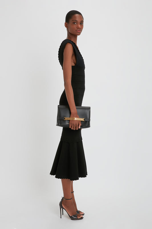 A person stands in profile view wearing a fitted black dress with a ruffled hem, black heels, and holding a chic Victoria Beckham Chain Pouch with Strap In Black Leather made of sleek Nappa leather.