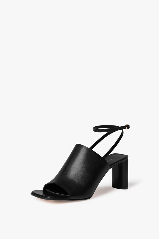 The Victoria Beckham Alina Sandal in Black is a single black open-toe, high-heel shoe with an adjustable ankle strap and a sculptural cylindrical heel, all set against a pristine white background.