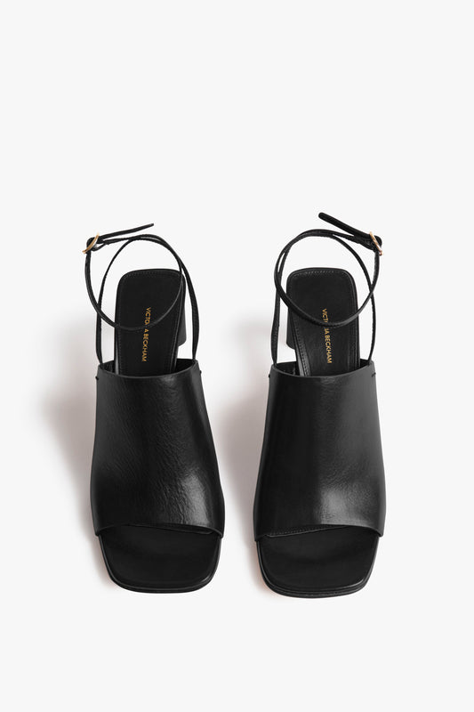 The Victoria Beckham Alina Sandal in Black is a pair of black open-toe slingback sandals with a sculptural block heel, featuring an adjustable ankle strap with a golden buckle.